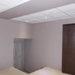 Wnętrze biura po odświeżeniu: ściany pomalowane na jasny fiolet, podwieszany sufit kasetonowy z wbudowanymi punktami świetlnymi, ciemne drzwi w prawym rogu i wejście do piwnicy po lewej stronie.