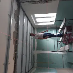 Pracownik na rusztowaniu, używający lasera krzyżowego do wyznaczania linii prostych na ścianie z zamontowanymi profilami pod płyty karton gips, widoczne płyty gipsowe i materiały budowlane