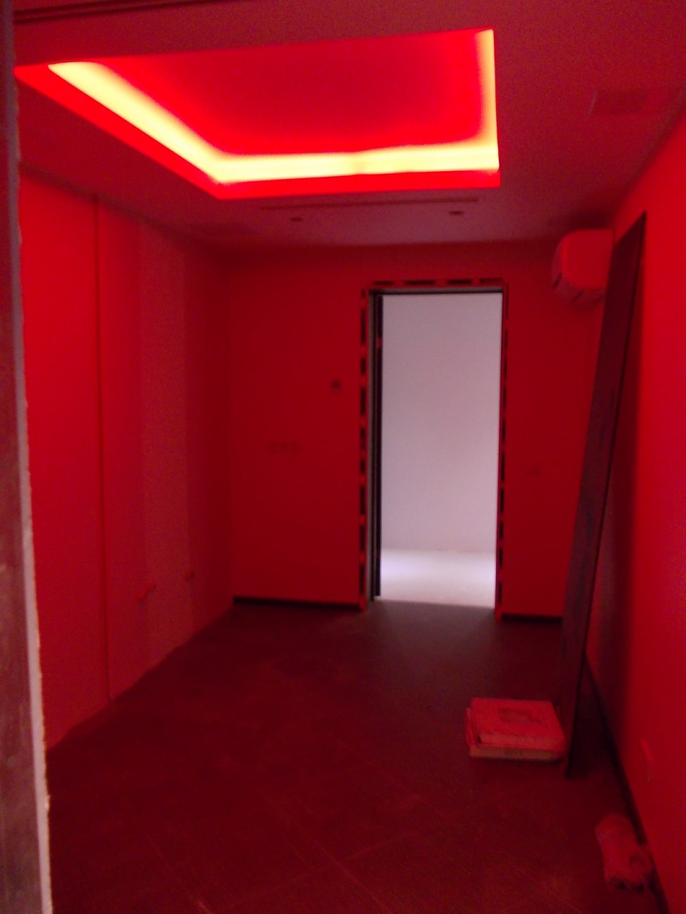 Wnętrze pokoju oświetlone intensywnym czerwonym światłem, z podświetlanym sufitem kasetonowym i otwartymi drzwiami do jaśniejszego pomieszczenia w tle. Na podłodze widoczne materiały budowlane.