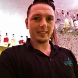 Uśmiechnięty barman w czarnej koszuli polo z logo na tle półki z butelkami w barze. Zdjęcie z dołu.