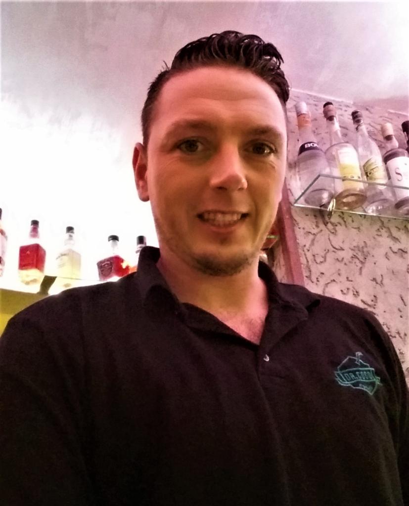 Uśmiechnięty barman w czarnej koszuli polo z logo na tle półki z butelkami w barze. Zdjęcie z dołu.