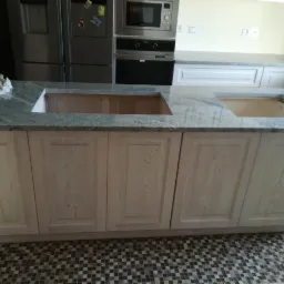 Wyspa kuchenna z szarym blatem kamiennym i jasnymi, drewnianymi szafkami, z widocznymi otworami na zlew i płytę grzewczą, butelka płynu na blacie, mozaikowa podłoga.