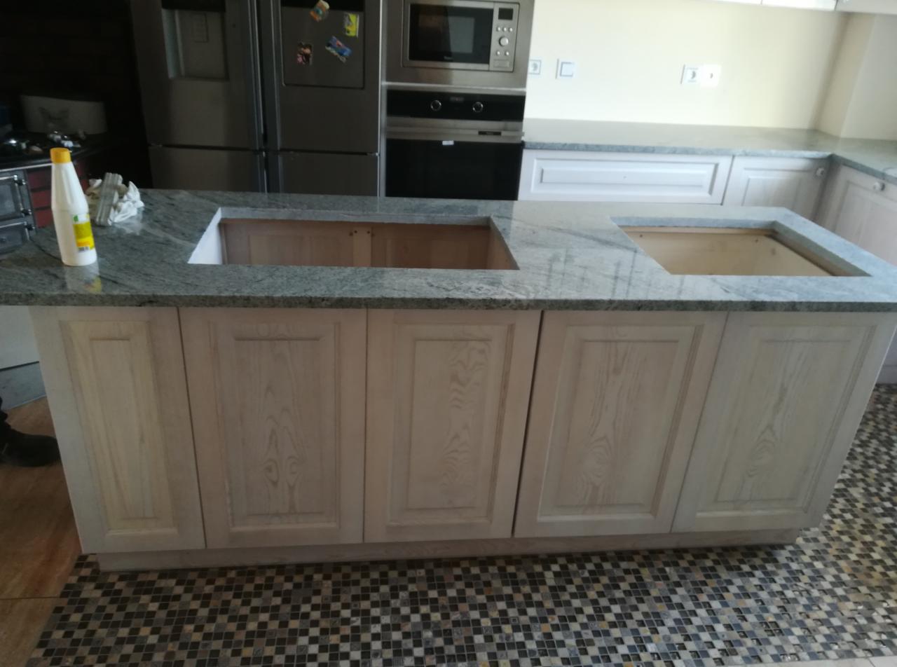 Wyspa kuchenna z szarym blatem kamiennym i jasnymi, drewnianymi szafkami, z widocznymi otworami na zlew i płytę grzewczą, butelka płynu na blacie, mozaikowa podłoga.