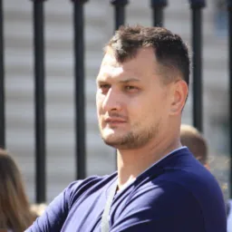 Mężczyzna z krótką fryzurą i lekkim zarostem, ubrany w granatową koszulkę, na tle metalowego ogrodzenia i rozmytych sylwetek innych osób.