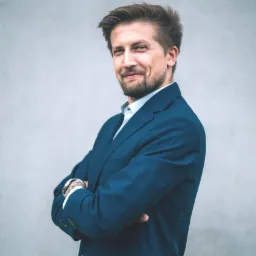 Uśmiechnięty mężczyzna w granatowym garniturze i białej koszuli, z tatuażem na nadgarstku, pozuje na tle szarej ściany. Profesjonalny wizerunek lektora.