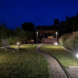 Oświetlony ogród nocą z altaną, ścieżką z kostki brukowej i lampami ogrodowymi, leżak w tle.