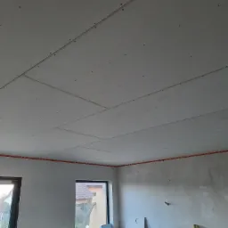 Montaż płyt gipsowo-kartonowych na suficie, widoczne wkręty mocujące, pomarańczowa listwa poziomująca przy ścianie, surowy stan wykończenia wnętrza.