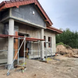 Dom jednorodzinny w trakcie prac elewacyjnych: widoczne rusztowania, ocieplenie ścian styropianem grafitowym oraz częściowo wykonane tynkowanie, czerwona dachówka.