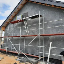 Budynek w trakcie ocieplania elewacji, widoczne szare płyty izolacyjne na ścianie, rusztowanie, poziomica oparta o rusztowanie, fragment dachu z czarną dach&oacute;wką, skrawki styropianu na ziemi.
