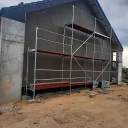 Dom w trakcie budowy z szarym tynkiem elewacyjnym, rusztowaniem i widocznymi bloczkami betonowymi, teren wokół w stanie surowym.