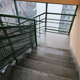 Betonowe schody w budynku z zieloną metalową barierką i oknem z widokiem na drzewa bez liści i rusztowania na budynku w pochmurny dzień.