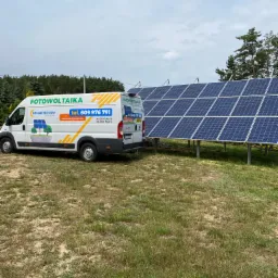 Gotowa instalacja fotowoltaiczna na trawiastym terenie obok lasu, z białym busem firmy Solar Tech PV zaparkowanym obok, na którym widnieje napis Fotowoltaika i numer telefonu.