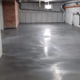 Świeżo wylana, gładka, szara posadzka w surowym pomieszczeniu z widocznymi instalacjami pod sufitem i fragmentem ceglanej ściany.