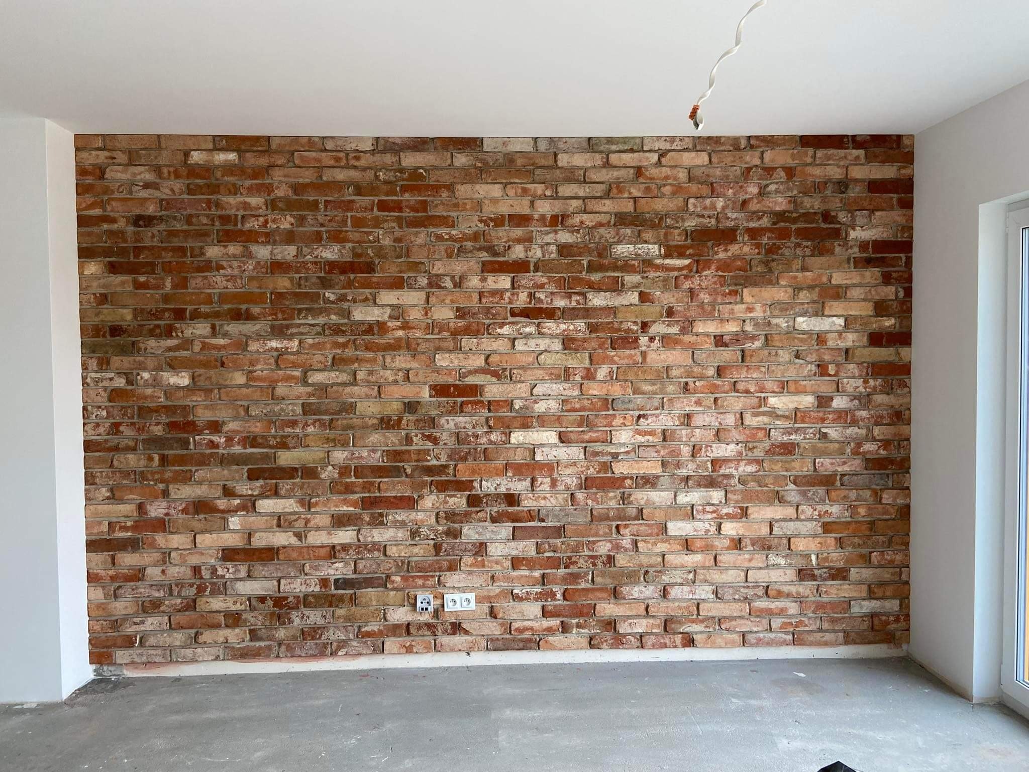 Ściana wewnętrzna wykończona cegłą klinkierową w odcieniach czerwieni i brązu, z widocznymi gniazdkami elektrycznymi i wystającym kablem z sufitu, w surowym, niewykończonym pomieszczeniu.