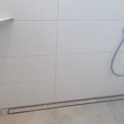 Wykończona ściana prysznica z białymi, prostokątnymi płytkami, odpływem liniowym w podłodze z szarych płytek i chromowanym wężem prysznicowym.