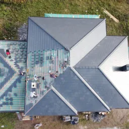 Widok z góry na budowę dachu: ekipa dekarzy układa ciemnoszare płytki na drewnianej konstrukcji, część dachu już pokryta, obok stoi biała przyczepa i leżą stosy drewna.