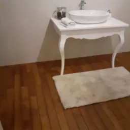 Biała umywalka nablatowa na ozdobnym stoliku z giętymi nogami, obok słoik z akcesoriami i zwinięty ręcznik, na podłodze beżowy dywanik, w tle jasne płytki ścienne i drewniana podłoga.