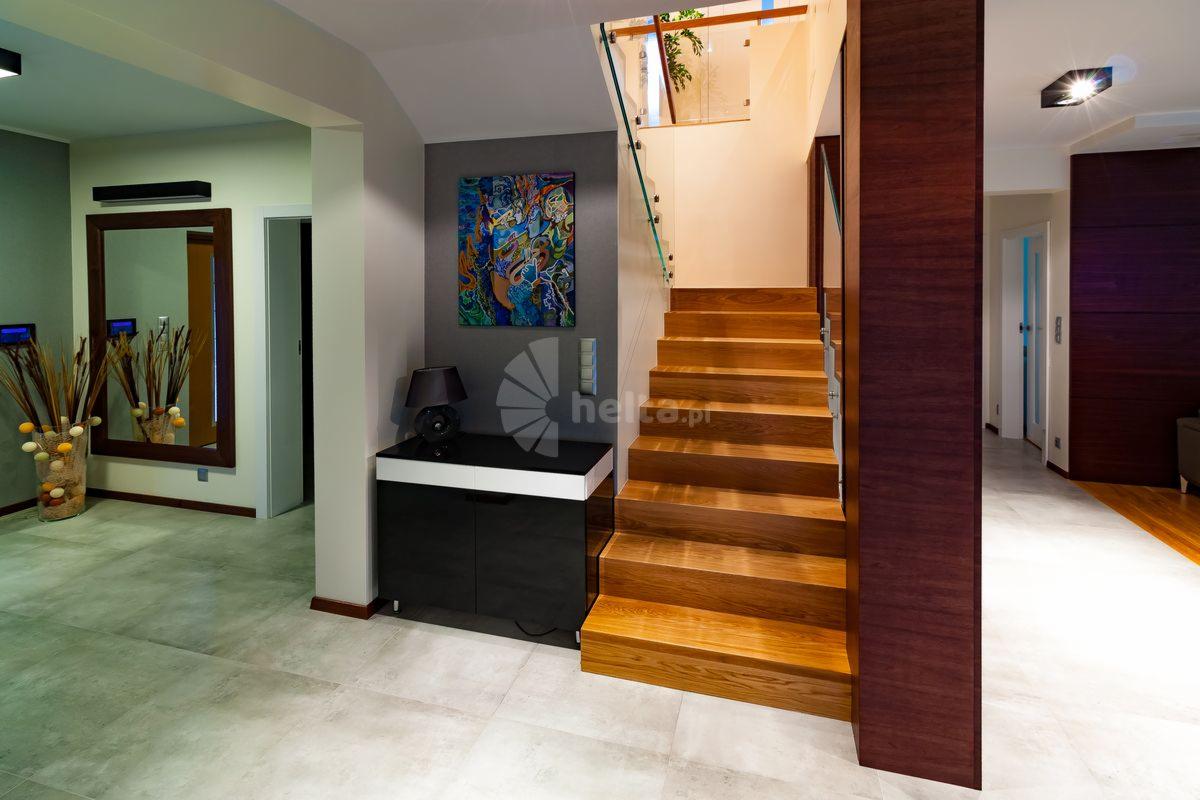 Nowoczesne schody drewniane z przeszkloną balustradą, oświetlone od góry, umieszczone w przestronnym holu z szarą ścianą i abstrakcyjnym obrazem.