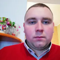 Portret mężczyzny w czerwonym swetrze na tle jasnego wnętrza z dekoracyjnym wazonem z kwiatami na półce. Neutralny wyraz twarzy, niebieskie oczy.