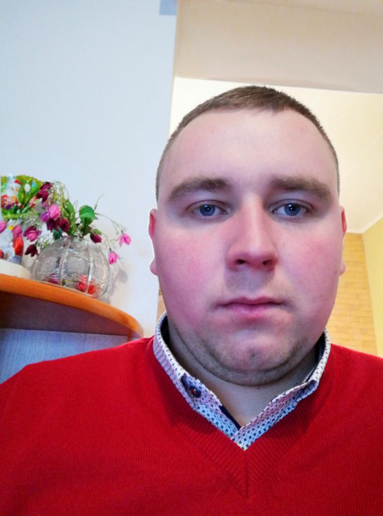 Portret mężczyzny w czerwonym swetrze na tle jasnego wnętrza z dekoracyjnym wazonem z kwiatami na półce. Neutralny wyraz twarzy, niebieskie oczy.