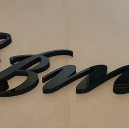 Czarny, błyszczący napis przestrzenny z elementem graficznym filiżanki kawy, umieszczony na jasnej, matowej powierzchni.