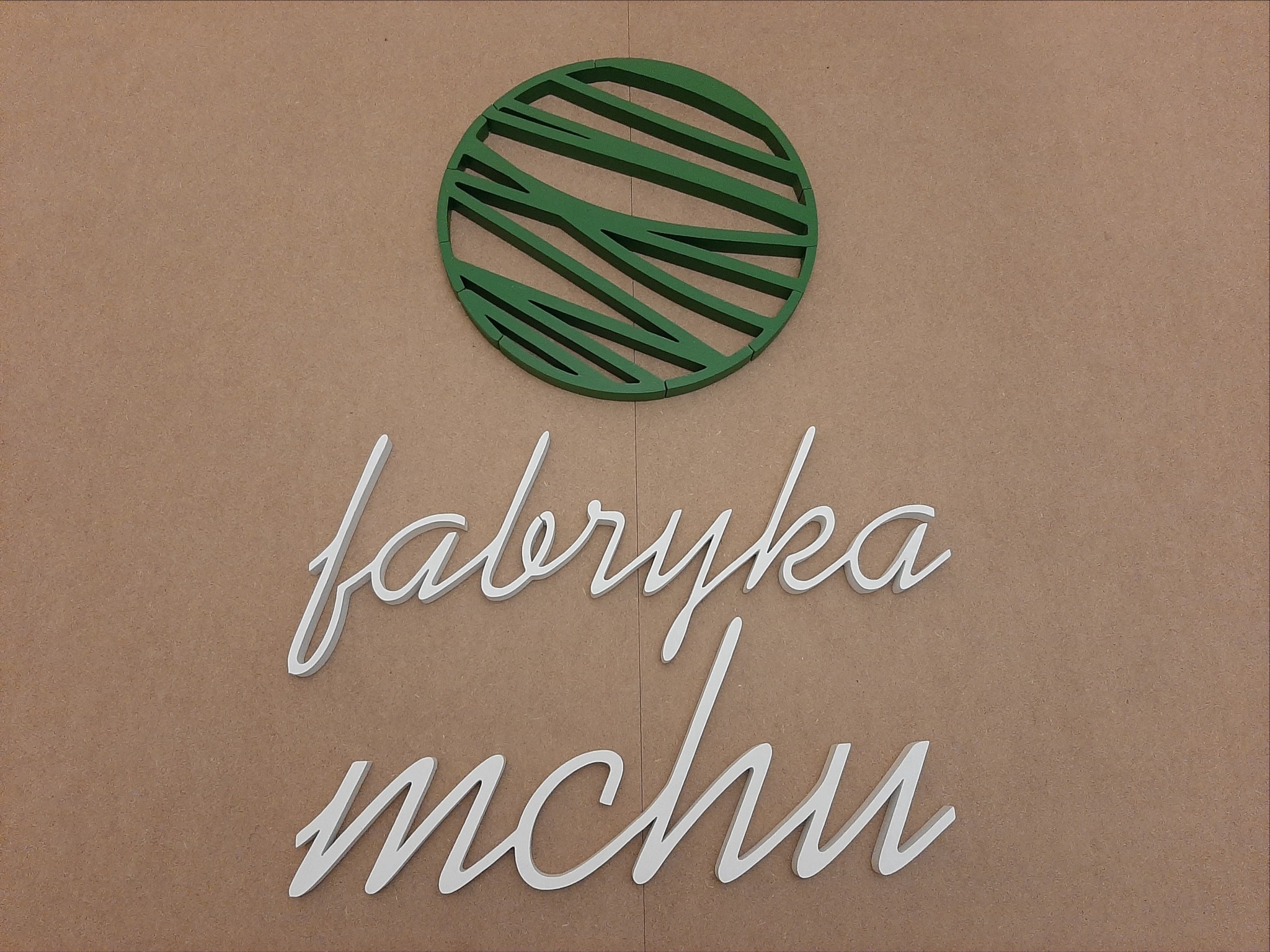 Logo firmy 'Fabryka Mchu' z zielonym, okrągłym symbolem przypominającym przekrój kuli ziemskiej zarysowany ukośnymi liniami na beżowym tle.