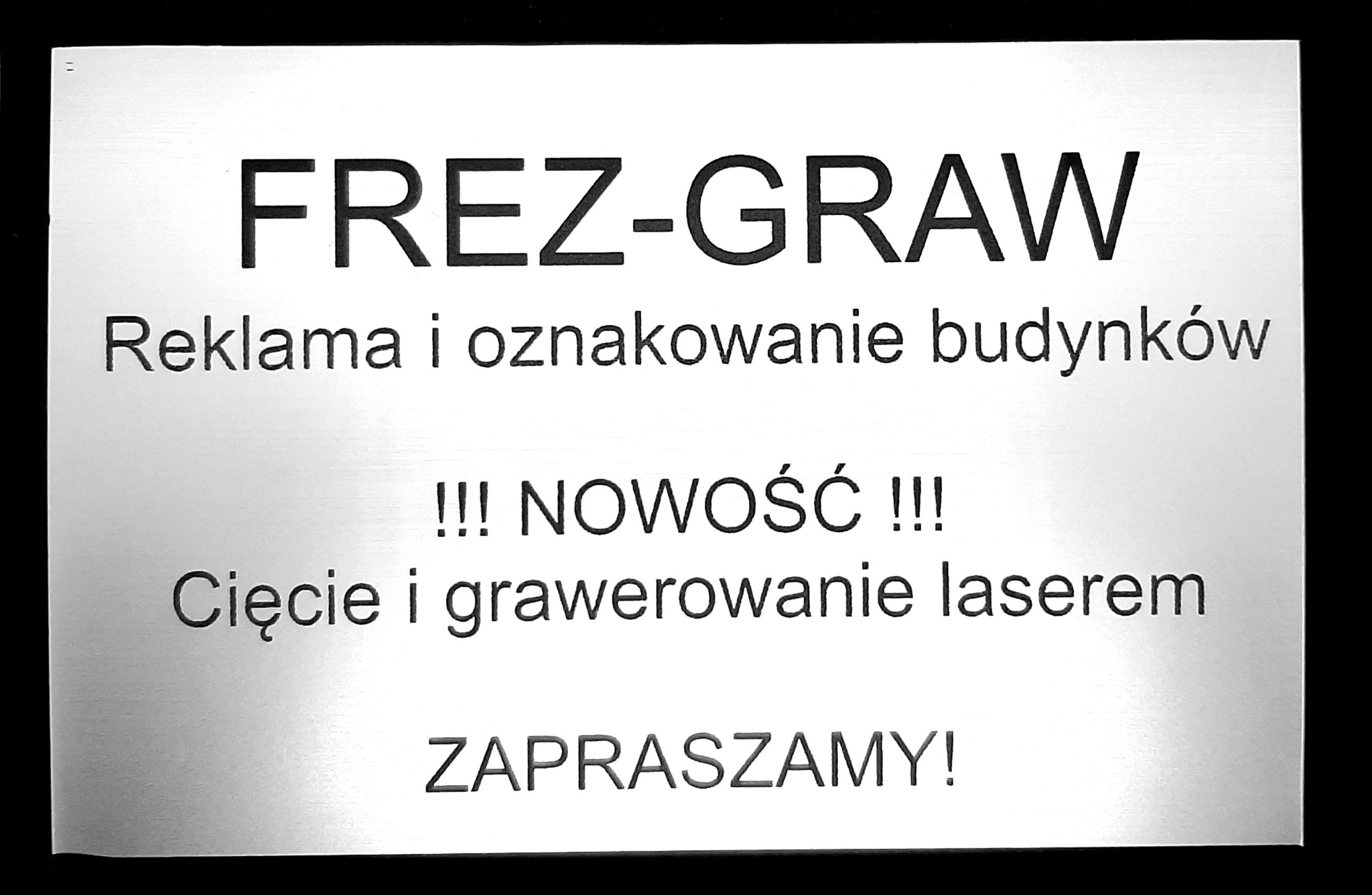 Srebrna tabliczka informacyjna z czarnym tekstem: FREZ-GRAW, Reklama i oznakowanie budynków, Nowość - cięcie i grawerowanie laserem, Zapraszamy.