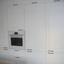 Biała zabudowa kuchenna z piekarnikiem w stylu klasycznym, czarne, poziome uchwyty, ściana wyłożona mozaiką w biało-czarną kratkę.