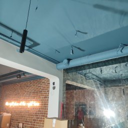 DD VENT - Niebieski sufit z widocznymi elementami instalacji wentylacyjnej, rurami i zawieszkami, z fragmentem ceglanej ściany z neonowym napisem w tle.