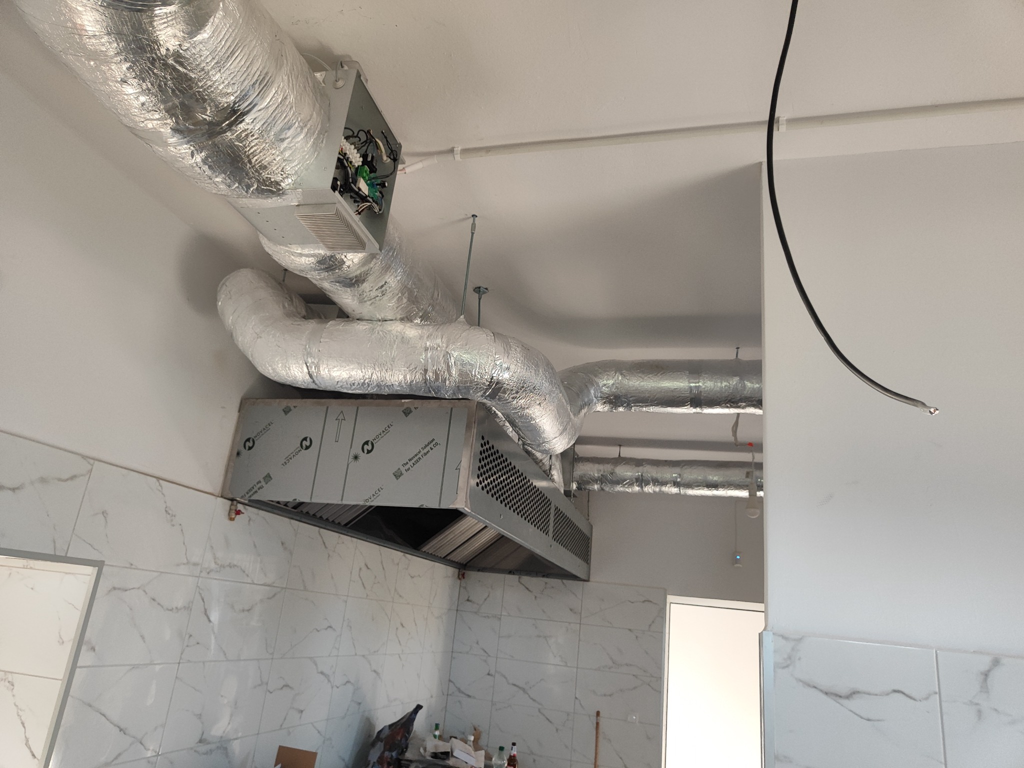 System kanałów wentylacyjnych z izolacją termiczną oraz metalowy okap z perforowaną obudową w pomieszczeniu wyłożonym białymi płytkami imitującymi marmur, widoczne elementy instalacji elektrycznej.