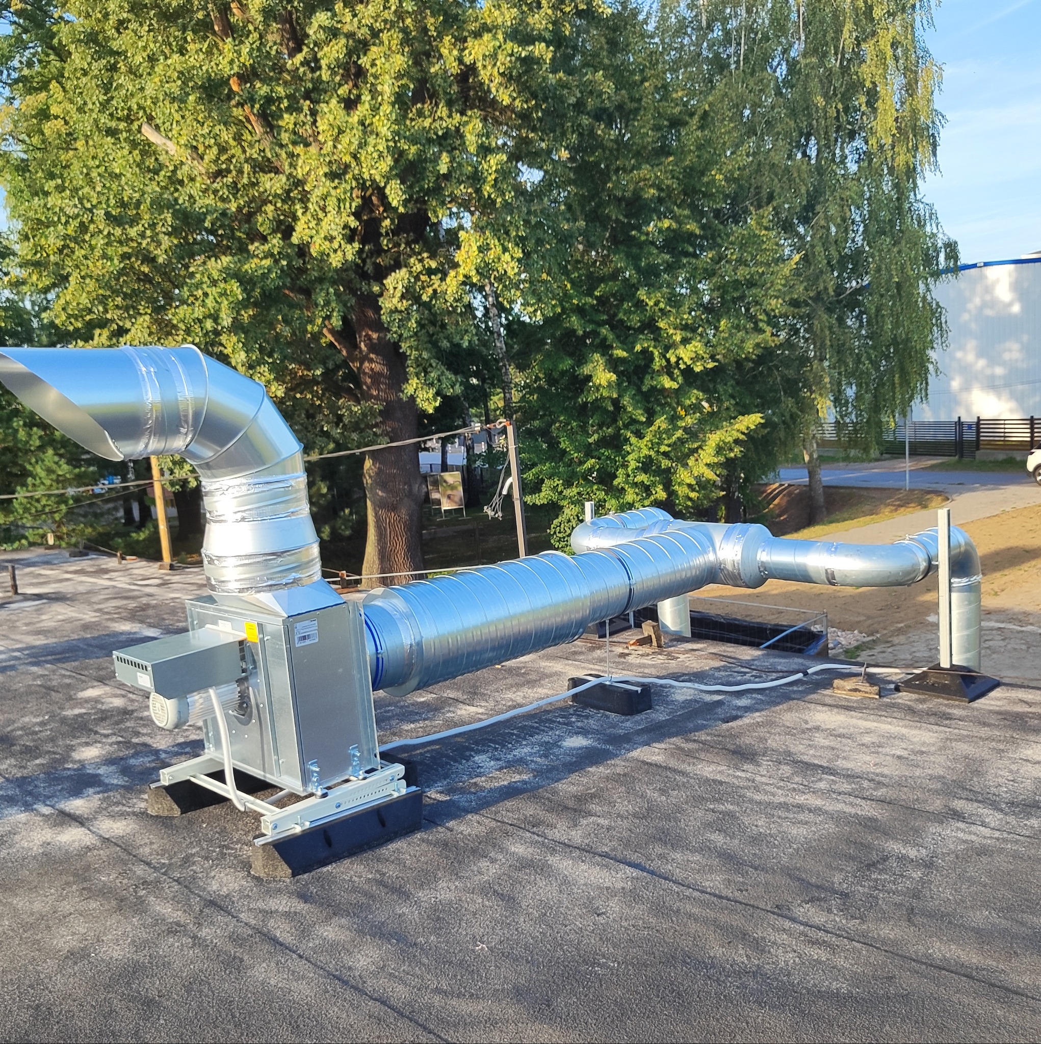 Kompletna instalacja wentylacyjna z centralą na płaskim dachu budynku komercyjnego, widoczne kanały wentylacyjne z blachy ocynkowanej, tło z drzewami i budynkiem.