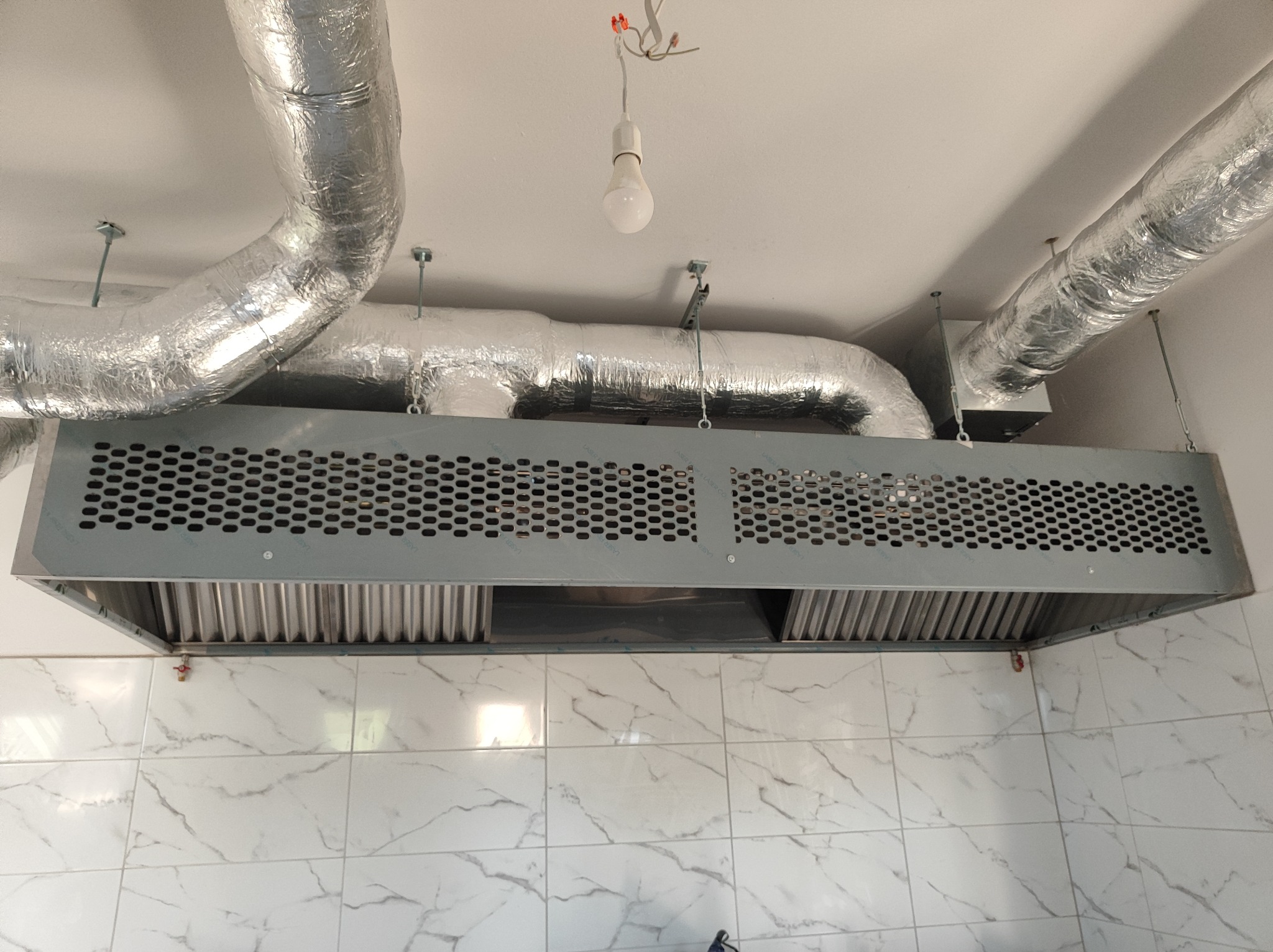 Srebrna, przemysłowa instalacja wyciągowa z perforowaną obudową i rurami wentylacyjnymi w folii aluminiowej, zamontowana pod sufitem w pomieszczeniu wyłożonym białymi płytkami imitującymi marmur.