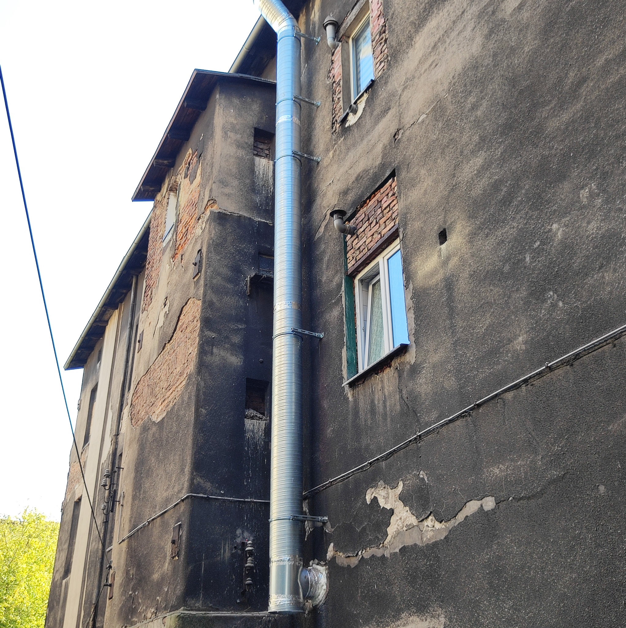Zewnętrzny, pionowy kanał wentylacyjny z blachy falistej, przymocowany do starej, zniszczonej ściany budynku z widocznymi ubytkami tynku i cegły.