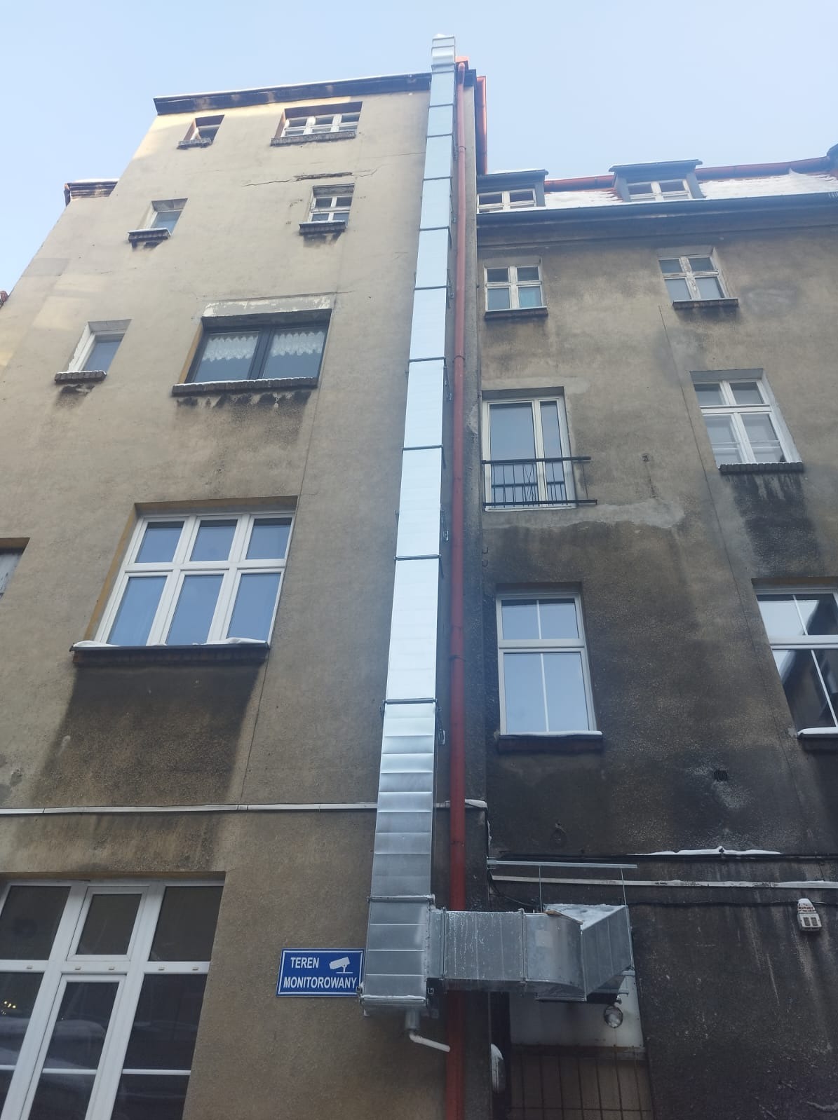 Zewnętrzny system wentylacyjny z blachy ocynkowanej, biegnący wzdłuż ściany kamienicy z widocznym znakiem 'Teren Monitorowany'.
