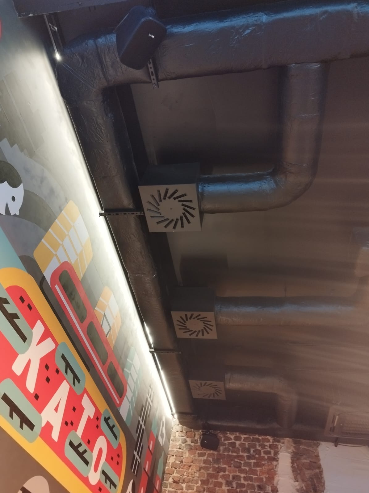 Szare, zaizolowane rury wentylacyjne pod sufitem z kwadratowymi nawiewnikami, widoczne fragmenty ściany z cegły i kolorowy mural.