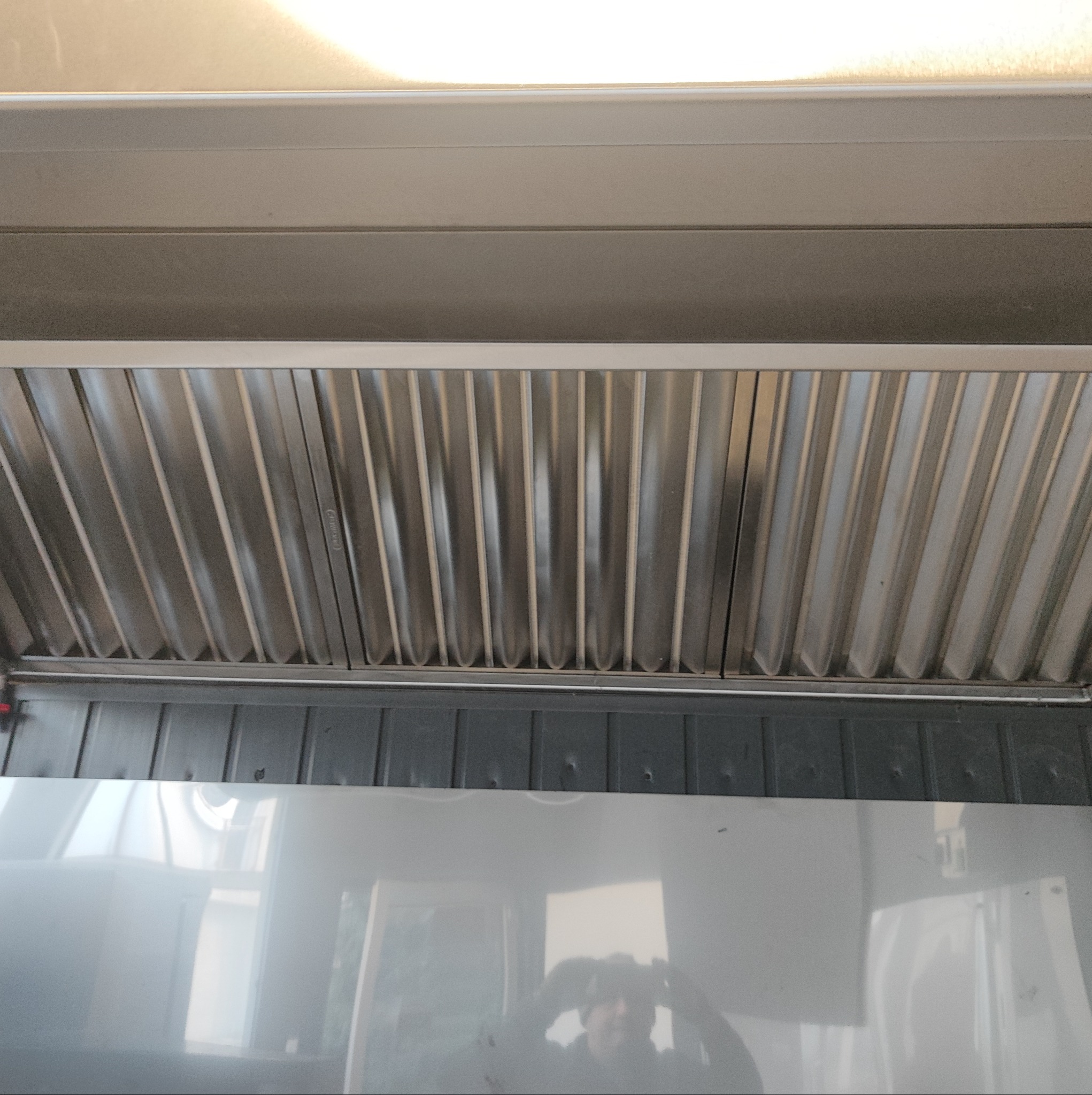 Wnętrze kanału wentylacyjnego z widocznym filtrem lamelkowym, zamontowanego pod sufitem, z odbiciem osoby robiącej zdjęcie na dolnej, błyszczącej powierzchni.