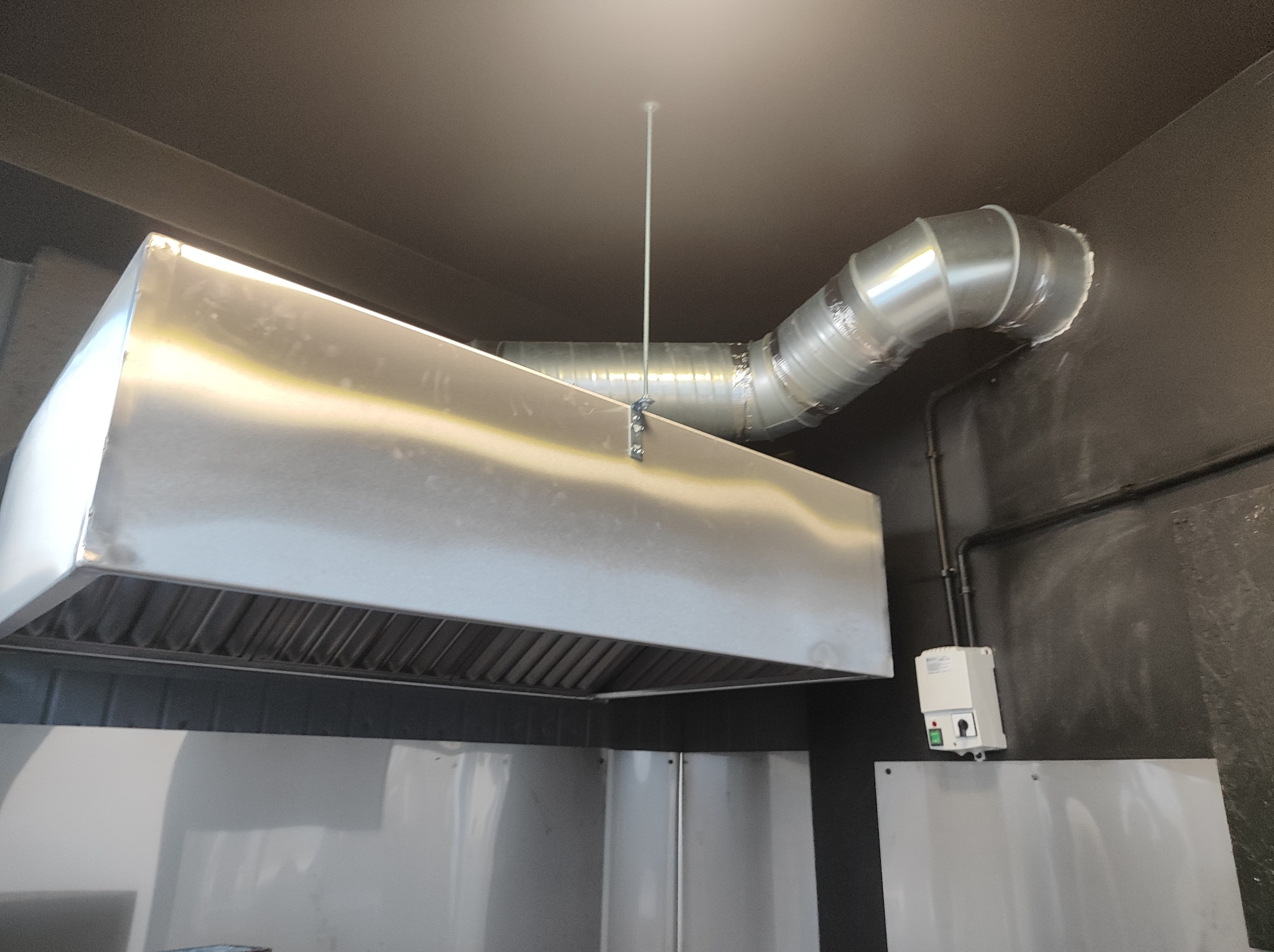 Srebrna, przemysłowa instalacja wentylacyjna z okrągłymi kanałami i prostokątnym okapem, zamontowana pod ciemnym sufitem w pomieszczeniu z panelami ściennymi, widoczny wyłącznik elektryczny.