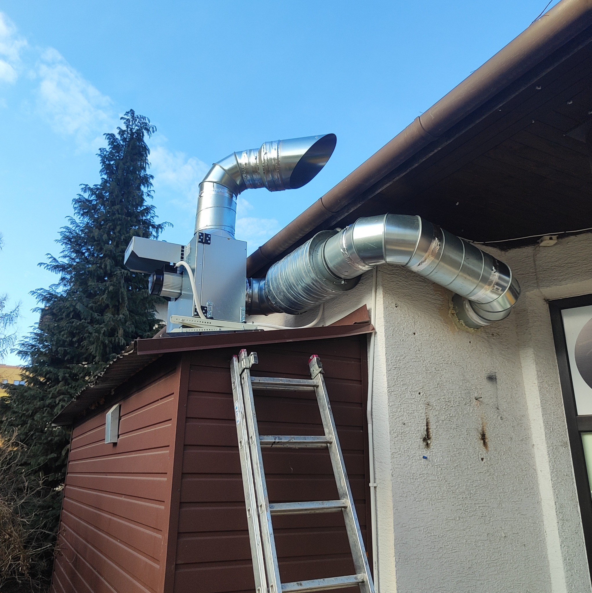 Zewnętrzny montaż systemu wentylacji z rurami spiro na budynku gospodarczym, widoczna drabina oparta o ścianę, błękitne niebo w tle.