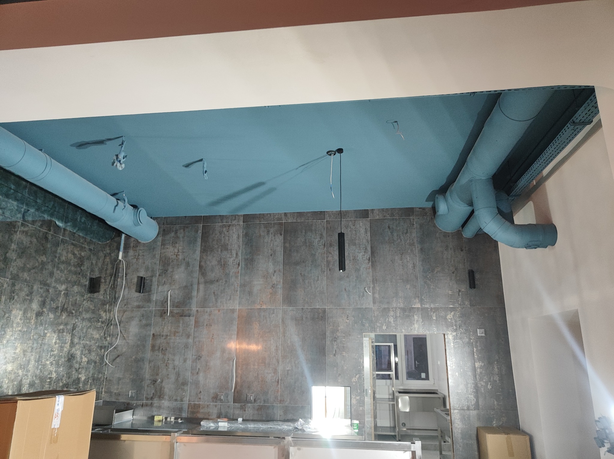 Niebieskie rury wentylacyjne zamontowane pod sufitem w pomieszczeniu z szarymi płytkami ściennymi, widoczna instalacja elektryczna i fragment wyposażenia kuchennego.