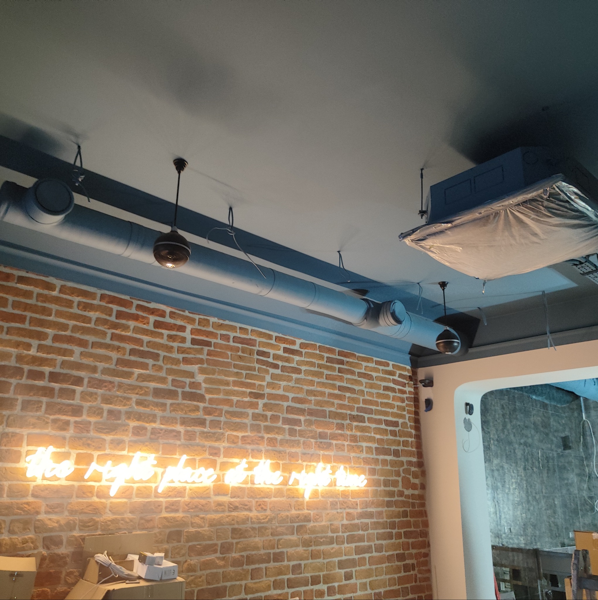 Instalacja wentylacji w lokalu komercyjnym z widocznymi rurami wentylacyjnymi, elementami oświetlenia i neonowym napisem na ceglanej ścianie.