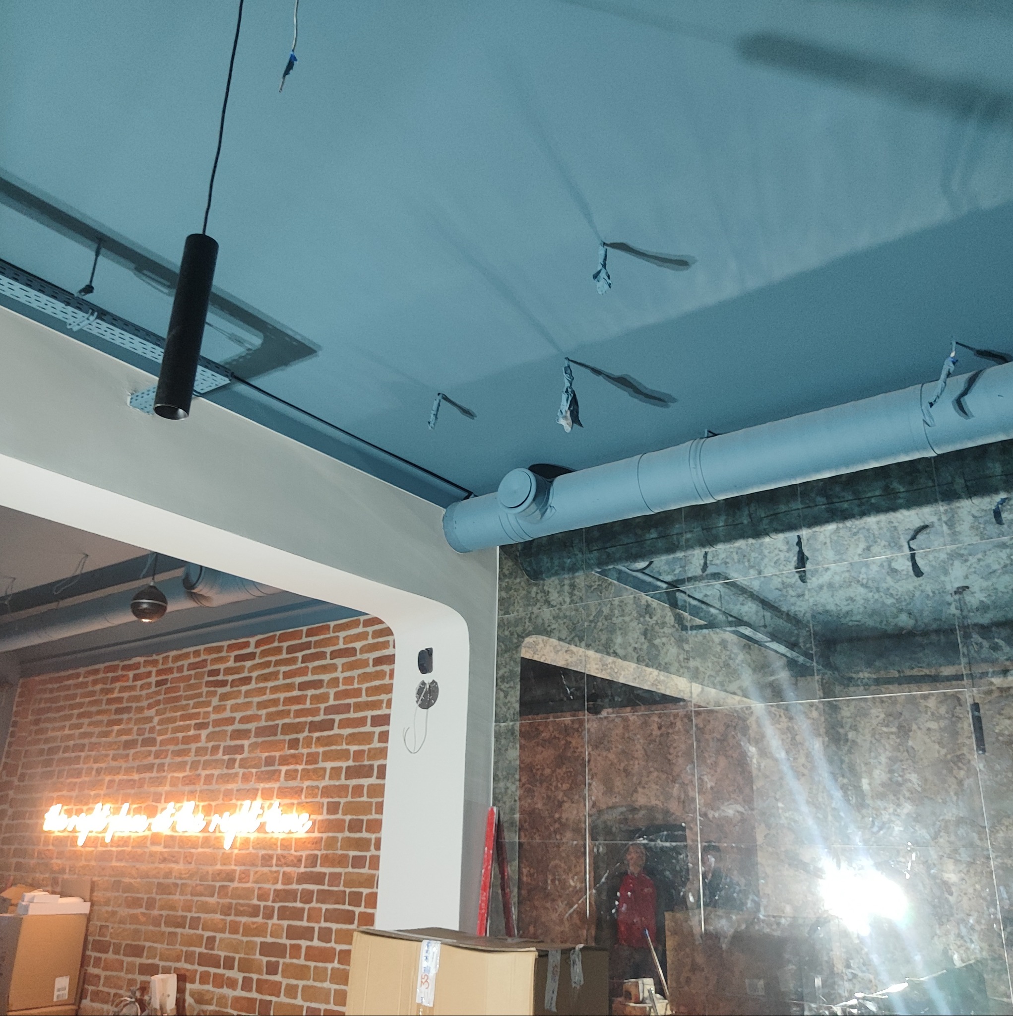 Niebieski sufit z widocznymi elementami instalacji wentylacyjnej, rurami i zawieszkami, z fragmentem ceglanej ściany z neonowym napisem w tle.