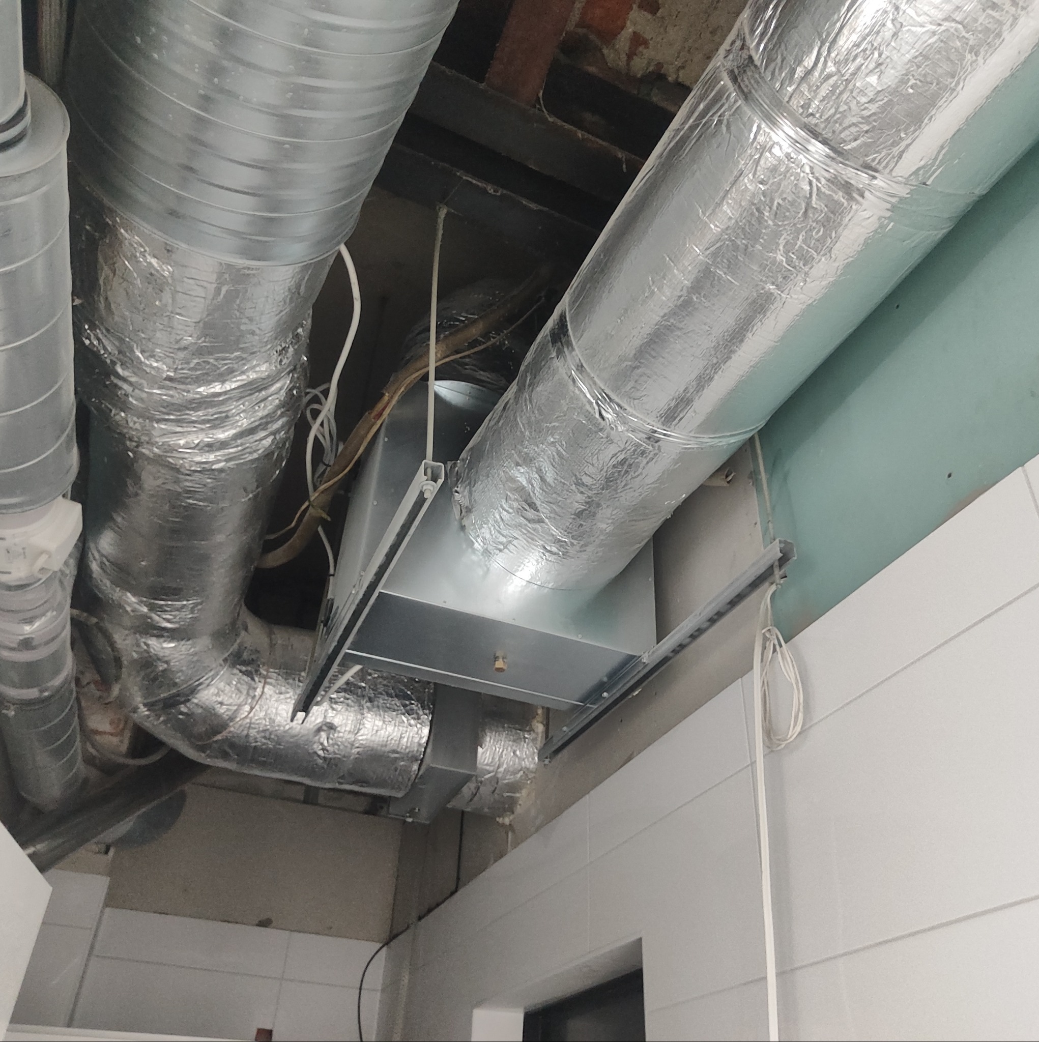 Fragment instalacji wentylacyjnej z aluminiowymi rurami i metalową skrzynką pod sufitem w pomieszczeniu z białymi panelami ściennymi.