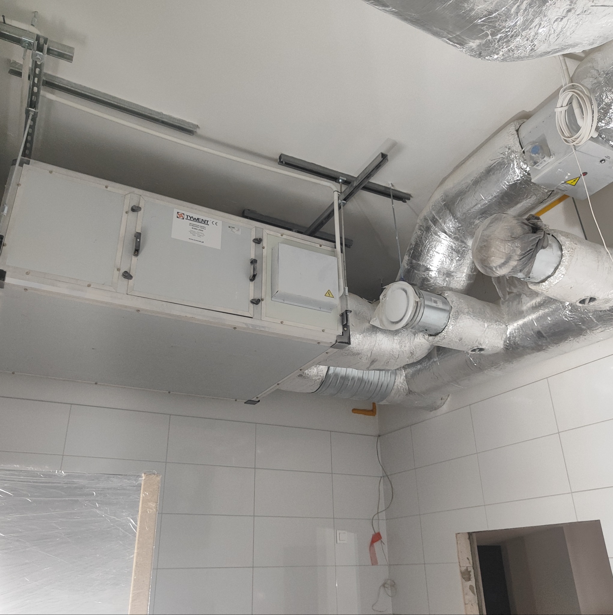 Centrala wentylacyjna marki Tywent zamontowana pod sufitem w pomieszczeniu z białymi kafelkami, widoczne kanały wentylacyjne w izolacji aluminiowej oraz elementy montażowe.
