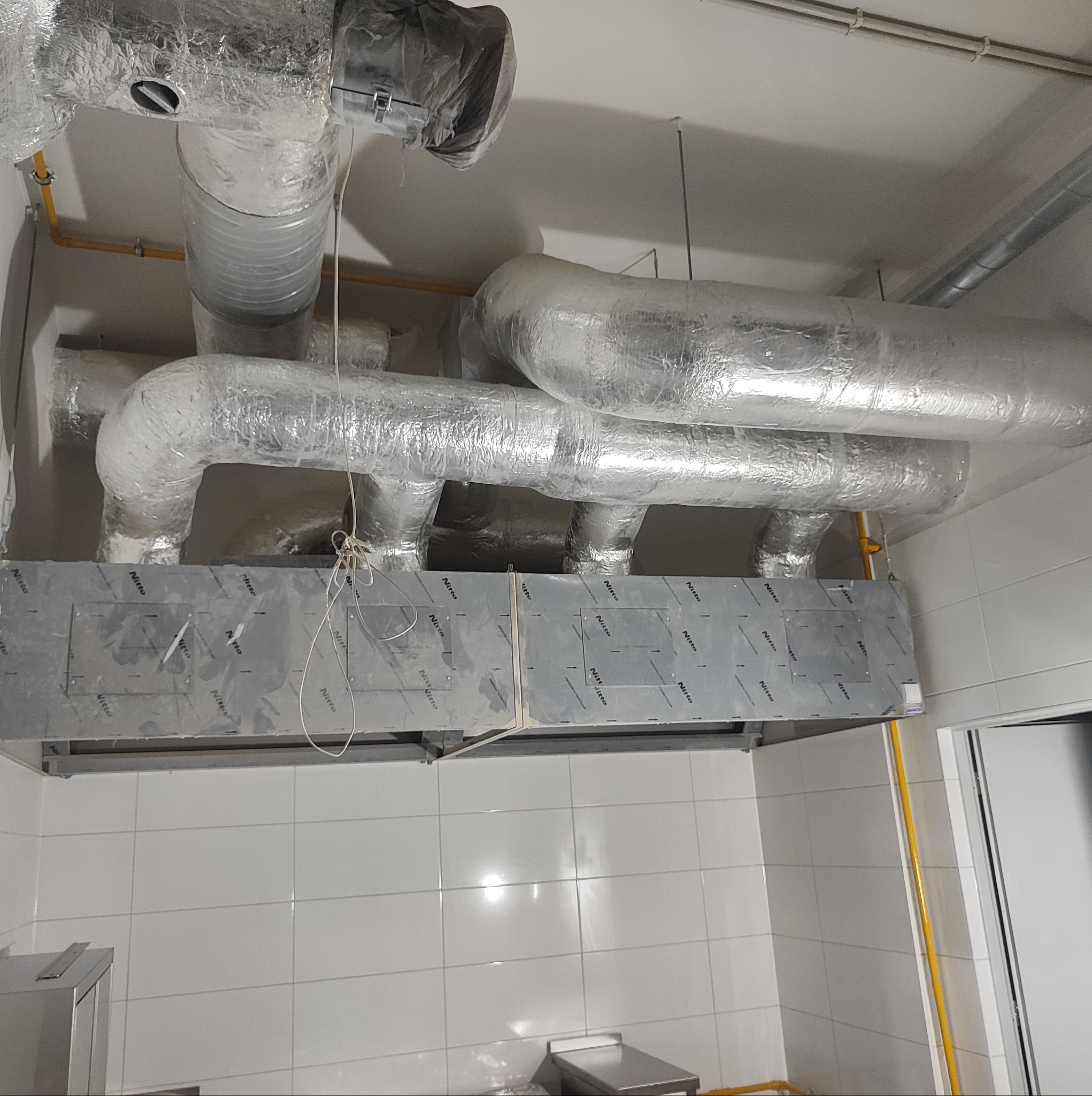 System kanałów wentylacyjnych z izolacją termiczną i centralą wentylacyjną z widoczną folią ochronną, zamontowany pod sufitem w pomieszczeniu wyłożonym białymi płytkami.