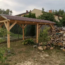 Nowa drewniana altana z brązowym dachem z gontu bitumicznego w ogrodzie, obok stos drewna opałowego, widok na dom z ceglanym kominem w tle.