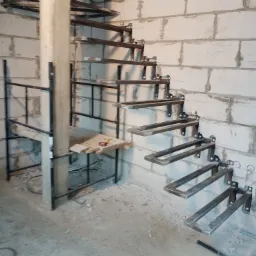 Wewnętrzne, metalowe schody spiralne w trakcie montażu w surowym stanie. Widoczne wsporniki przymocowane do ściany z białych bloczków, tymczasowe rusztowanie i narzędzia.