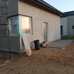 Budynek w trakcie prac wykończeniowych elewacji zewnętrznej, okna zabezpieczone folią, widoczne rusztowanie i narzędzia.