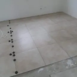 Układanie beżowych płytek podłogowych z użyciem systemu poziomowania; widoczne czarne klipsy i kliny, a także fragment nieukończonej podłogi.