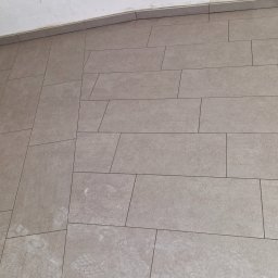Artur Wolski usługi remontowo budowlane - Ułożona podłoga z prostokątnych, beżowych płytek ceramicznych z ciemniejszą fugą, widoczne ślady stóp i fragment schodka z wykończeniem.