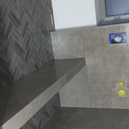 Artur Wolski usługi remontowo budowlane - Szara łazienka z płytkami ułożonymi w jodełkę na ścianie, półka z płytek, otwory kanalizacyjne w ścianie, okno z szarą ramą.